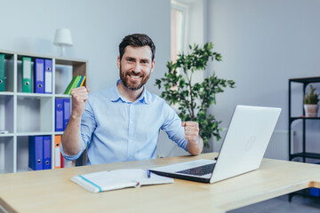 Successful businessman with a beard looks at the camera and smiles, celebrates triumph, works on a laptop in the office