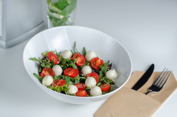 Caprese salad with fresh cherry tomatoes, mini mozzarella, pesto, basil, olive oil and balsamic sauce, with lettuce and arugula on white plate. A plate of salad on a white table in a restaurant.