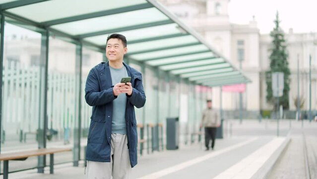 Senior Chinese tourist uses mobile phone app to book accommodation and call taxi, man near airport with suitcase. Asian man using smartphone app to rent a car at the bus stop. waiting public transport