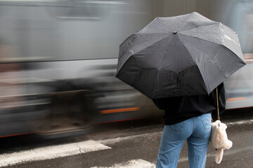 Young woman in jeans under black umbrella on a crossroad on a rainy day with traffic in motion blur,  ear view - 501621902