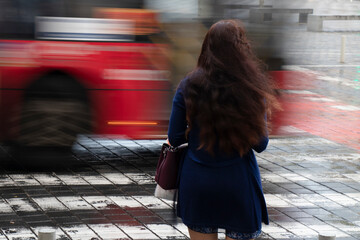 Young woman with long wavy hair waiting on a crossroad on a rainy day with traffic in motion blur, rear view - 501621905