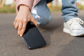 Kid girl picking damaged mobile phone with cracked screen from asphalt outdoors, closeup