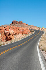 Overton, Nevada, USA - February 24, 2010: Valley of Fire. Closeup of bended black asphalt road with yellow divider under blue sky bordered by red rocks. Yellow traffic sign far off.