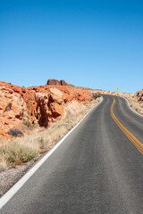 Overton, Nevada, USA - February 24, 2010: Valley of Fire. Bended black asphalt road with yellow divider under blue sky bordered by red rocks. Yellow traffic sign far off.