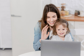 Happy young woman with joyful toddler having video call from relatives, waving hello to them. Beautiful mother with little daughter smiling, using computer to talk online with family while quarantine