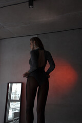 young teen girl in a shiny silver bodysuit stands fashion from the back with hands near waist on the gray art wall background in loft interior with red light and mirror. fashion concept, free space