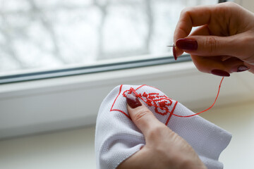 Hands of a girl, woman sew a small embroidery pattern on a white canvas. The ornament is made with...