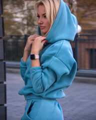 teen leisure. young blonde girl in blue oversize costume with hoodie  stands casual with hands near hood and looks apart with cute smile on the street park background. lifestyle concept, free space