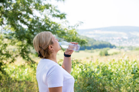 Profile photo of a sporty blonde woman wearing digital watch on her hand and is drinking water from the plastic bottle outdoors after jogging.