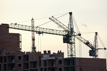 Dark silhouette of tower cranes at high residential apartment buildings construction site at sunset. Real estate development