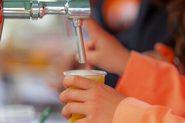 Selective focus of hand holding a plastic glass beer under draught beer tap, Outdoor party celebration of the birthday of the King in Netherlands, National holiday King’s Day or Koningsdag in Dutch.