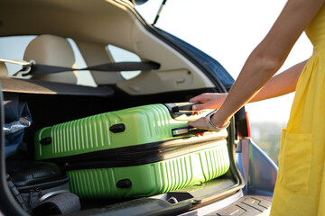 Young woman in yellow summer dress taking green suitcase from car trunk. Travel and vacations...