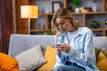 Amazed woman watching media content in a smart phone sitting on a sofa in the living room in a house interior