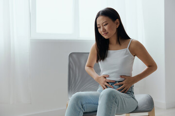 Fototapeta na wymiar Stomach ulcer Gastritis. Tormented suffering tanned beautiful young Asian woman hold hands on stomach at home interior living room. Injuries Poor health Illness concept. Cool offer Banner