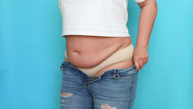 Unrecognizable fat plump overweight woman wearing beige underwear, white T-shirt, pulling on blue jeans, buttoning up, adjusting on blue background. Body positive, obesity, weight loss, liposuction.