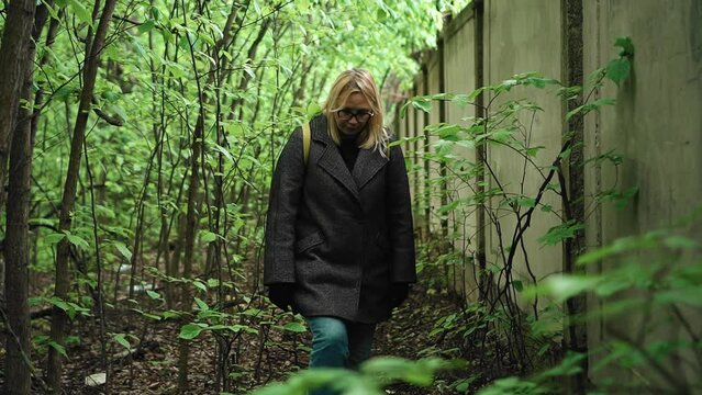 A girl in glasses and a gray coat moves along the wall through thickets of grass and trees. Shooting from the front in motion