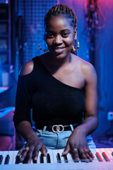 Vertical medium portrait of young Black woman with digital keyboard in recording studio smiling at camera