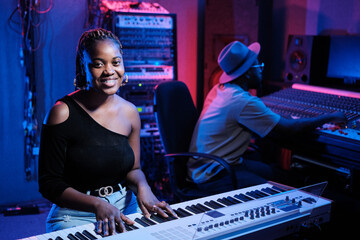 Joyful young African American female musician playing digital keyboard while producer working on mixing console in recording studio