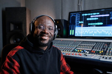Portrait of cheerful African American music producer wearing headphones and eyeglasses sitting in recording studio smiling at camera