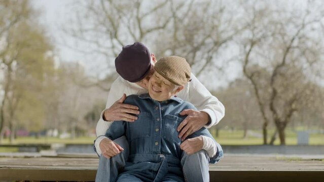 Adorable elderly couple enjoying sunny day in park together. White-haired senior man kissing his wife while couple sitting at parapet and having good time outdoors. Love, retirement concept