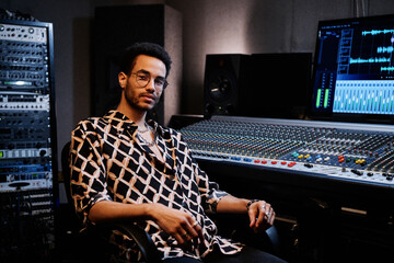 Young African American recording producer wearing fashionable outfit sitting against mixing console...