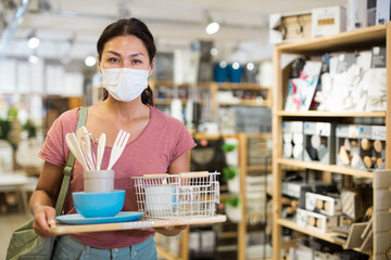 Asian woman customer in face mask choosing tableware and kitchen accessories at household goods...