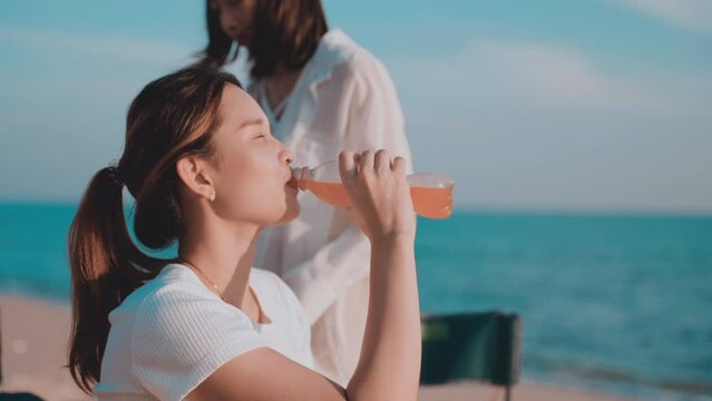 Slow motion footage of Asian woman attractive sitting on picnic chair holding orange juice bottle and drinking on the beach in vacation summer. Young female and her friend drink cocktail at seaside. 