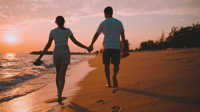 Slow motion of happy Asian couple hands holding and walking together on the beach while golden sunset time evening of summer vacation. Young in love romantic and relaxing at seaside. Honeymoon concept