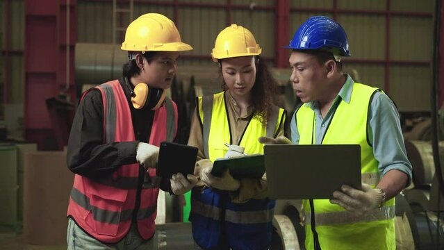 Team of professional Asian workers wears safety hard hats holding laptop, tablet, clipboard with meeting, talk, discuss plan in factory industry. Production leader standing training for employees. 