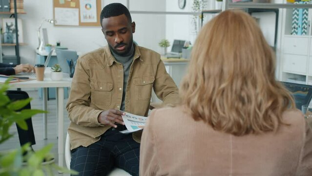 Slow motion of business consultant talking to client indoors in modern office. African American man holding papers sharing ideas with Caucasian woman.