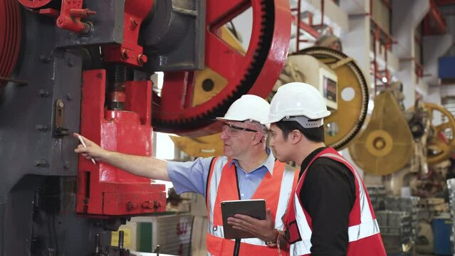 Senior man engineer wearing safety goggles and hard hat standing holding tablet with training young engineer operate hydraulic press stamp machine in factory. Two industry maintenance workers working.
