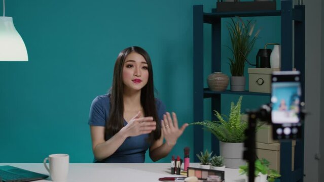 Asian woman filming makeup tutorial with smartphone camera, giving beauty tips for social media audience. Online blogger recording video with cosmetics, eyeshadow palette and brush.
