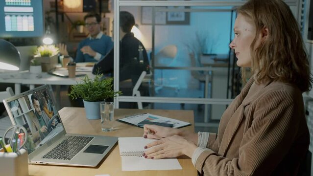 Ambitious businesswoman talking to male colleague making video call using laptop and earpods in office at night. People and communication at night.