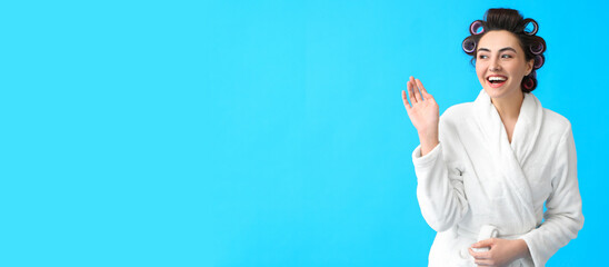 Beautiful young woman in bathrobe and hair curlers on blue background with space for text - obrazy, fototapety, plakaty