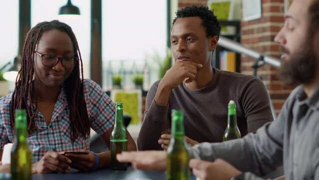 Portrait of african american man playing board games with cards and serving alcoholic beer in bottle. Happy people gathering together at home to play fun activity game with tactics.