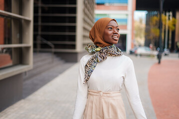 Smiling Muslim black woman in hijab standing near office building - obrazy, fototapety, plakaty
