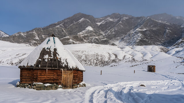 The old wooden hexagonal Altai house - ail in a snow-covered valley. Icicles hang from the conical roof. A well-trodden path winds through snowdrifts. A mountain range against a blue sky. 