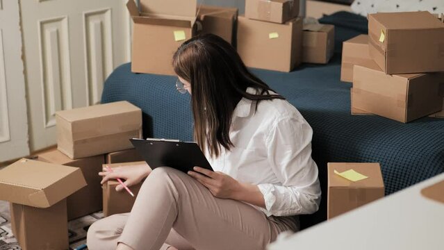 Woman sitting on floor at home, counting and marking boxes in tablet, works on an online marketplace, selling goods. There are many boxes around. Sells goods of own production on online platforms.
