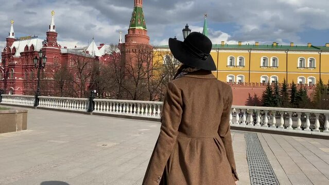 Young beautiful lady elegance madame in hat and coat walking on Red Square  near Historical Museum, Kremlin, Alexander Garden and Okhotny Ryad. Spring, blooming flowers. Center of Moscow, Russia.