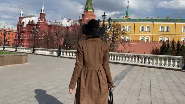 Young beautiful lady elegance madame in hat and coat walking on Red Square  near Historical Museum, Kremlin, Alexander Garden and Okhotny Ryad. Spring, blooming flowers. Center of Moscow, Russia.