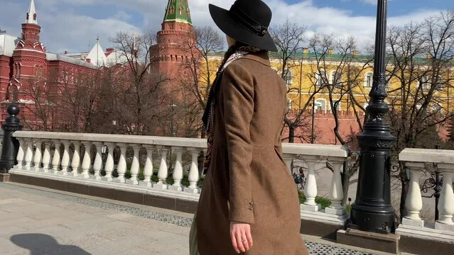Young beautiful lady elegance madame in hat and coat walking on Red Square  near Historical Museum, Kremlin, Alexander Garden and Okhotny Ryad. Spring, blooming flowers. Center of Moscow, Russia.