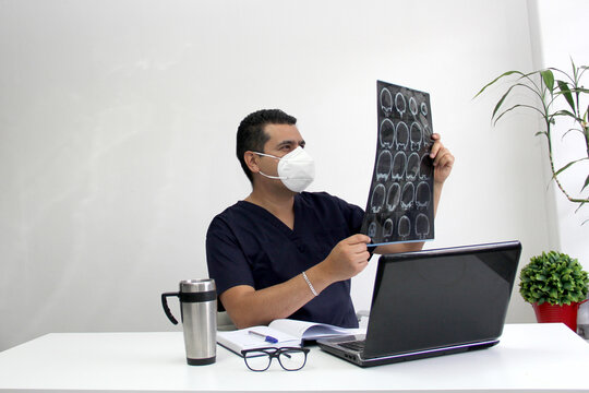 Latino neurologist doctor man with face mask gives remote consultation in the new normality due to the Coronavirus pandemic analyzes a brain tomography
