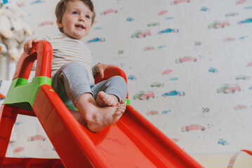 Happy little 2 years boy rides on a slide in the children's room and plays with toys. Kindergarten for toddlers. Blurred and unfocus image