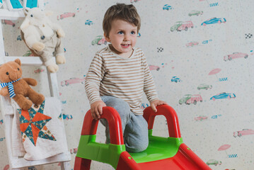 Happy little 2 years boy rides on a slide in the children's room and plays with toys. Kindergarten...