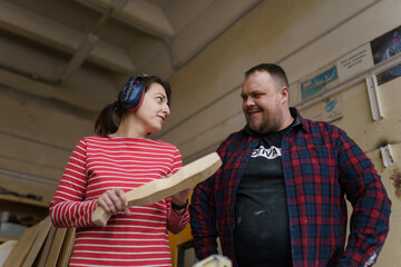 A female carpenter in a striped red shirt and headphones communicates with a male carpenter in a plaid shirt, standing in a wood workshop