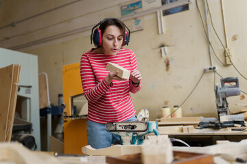 A female carpenter in a red-striped shirt stands in her workshop, plumes untreated wood and blows dust off a wooden plank