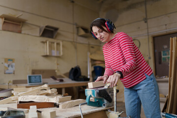 A carpenter woman in a red striped shirt grinding raw wood with ander tool in her workshop