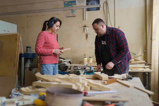 A female carpenter in a striped red shirt and headphones communicates with a male carpenter in a plaid shirt, standing in a wood workshop - Powered by Adobe