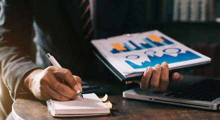 Male businessman use a laptop computer and take notes in the notebook at the office desk in the...
