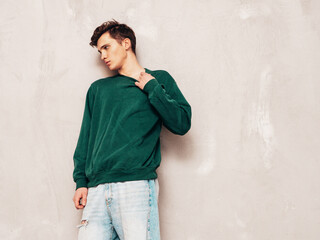 Portrait of handsome confident stylish hipster lambersexual model.Man dressed in green sweater and jeans. Fashion male posing in studio near grey wall